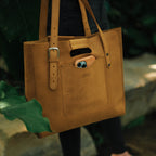 Everyday Leather Tote in Tobacco carried outdoors among leafy plants, with a phone in the front pocket