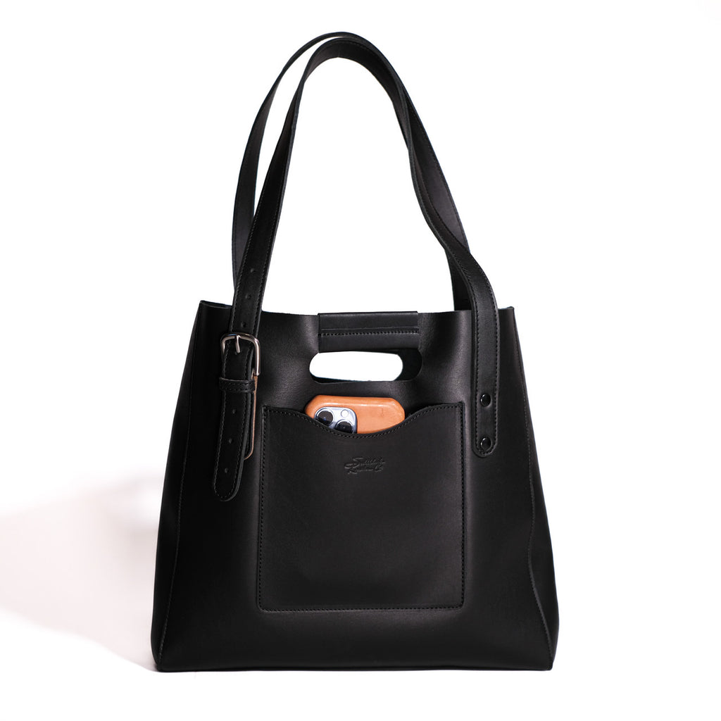 Everyday Leather Tote in Black, front view with shoulder handles, buckled side strap and a phone in the front pocket