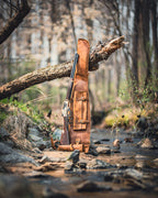 Shotgun Sleeve in Tobacco standing upright by a forest creek beside a lever-action rifle