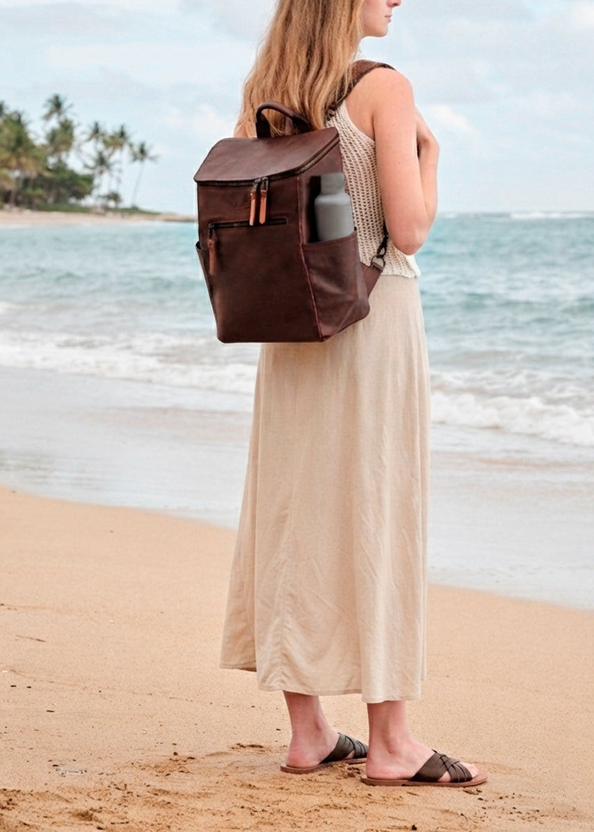 All in One Leather Backpack in Chestnut worn by a woman standing on a sandy beach facing the ocean