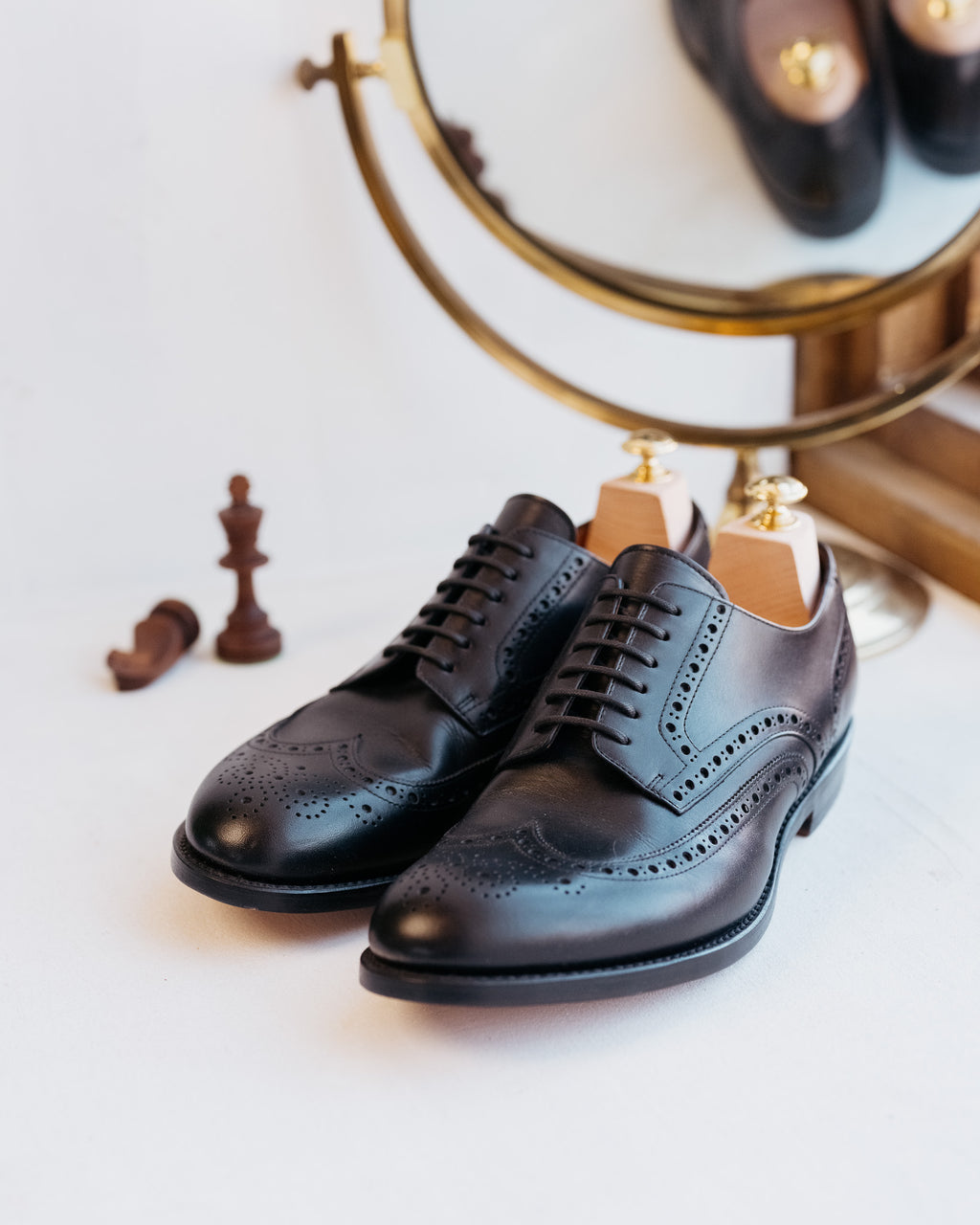Wingtip Shoes in Raven Black, pair with shoe trees beside a brass mirror and chess pieces