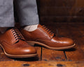 Wingtip Shoes in Natural Tan, worn with gray trousers on a rustic wood floor