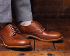 Wingtip Shoes in Natural Tan, worn with gray trousers on a rustic wood floor