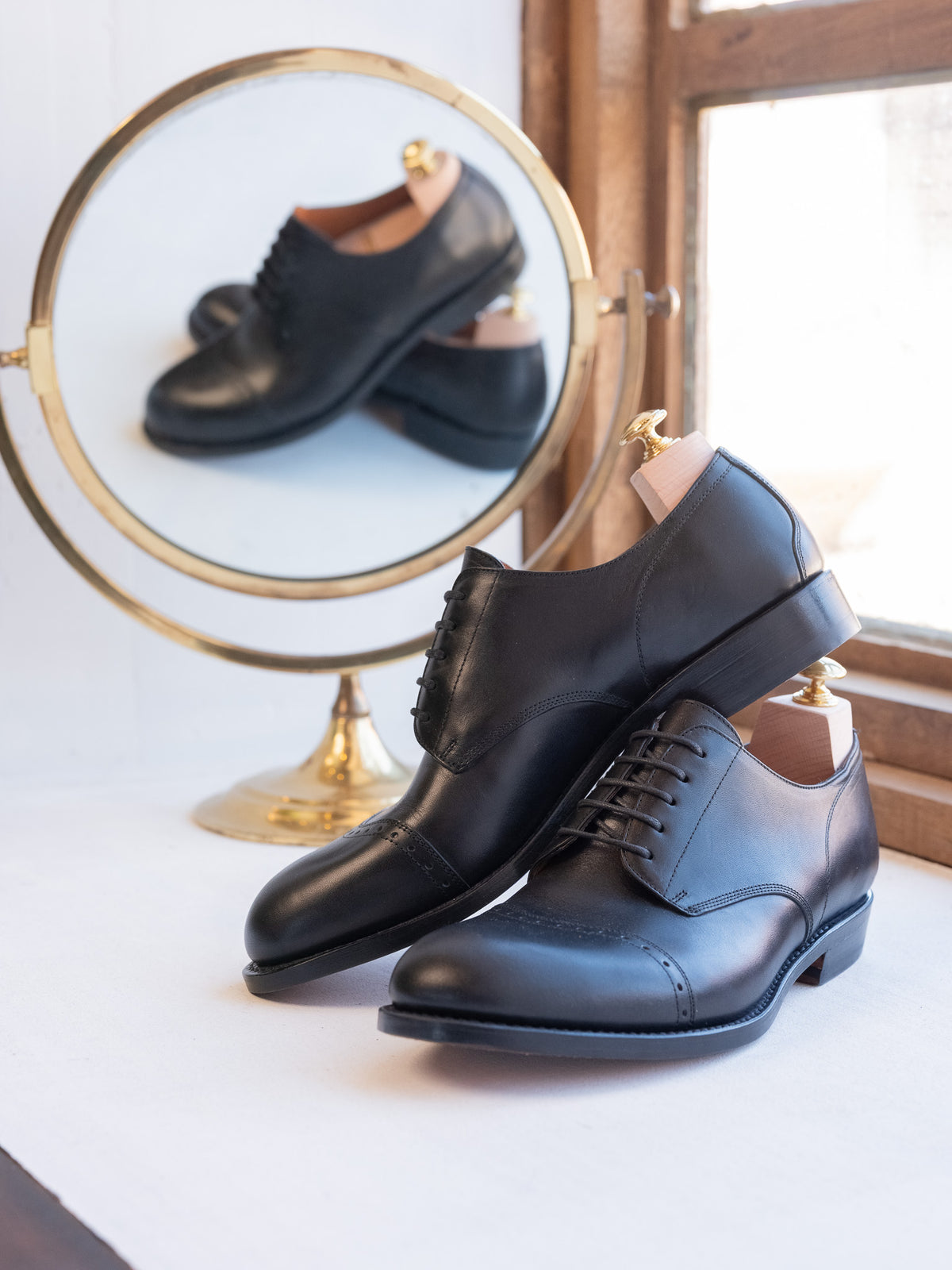 Toe Cap Shoes in Raven Black, pair on shoe trees beside a round brass mirror by a window
