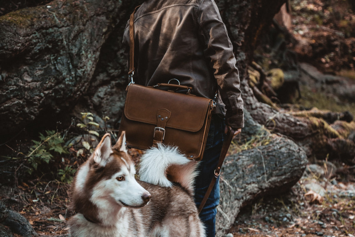 Slim Leather Briefcase in Chestnut carried by a person in a leather jacket on a forest trail beside a husky