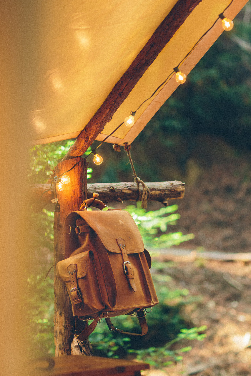 Tank Backpack in Tobacco, hanging from a wooden post under a canopy with string lights in the forest