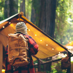 20" Waterbag in Tan, carried backpack style by a hiker beside a lit canvas tent in the forest