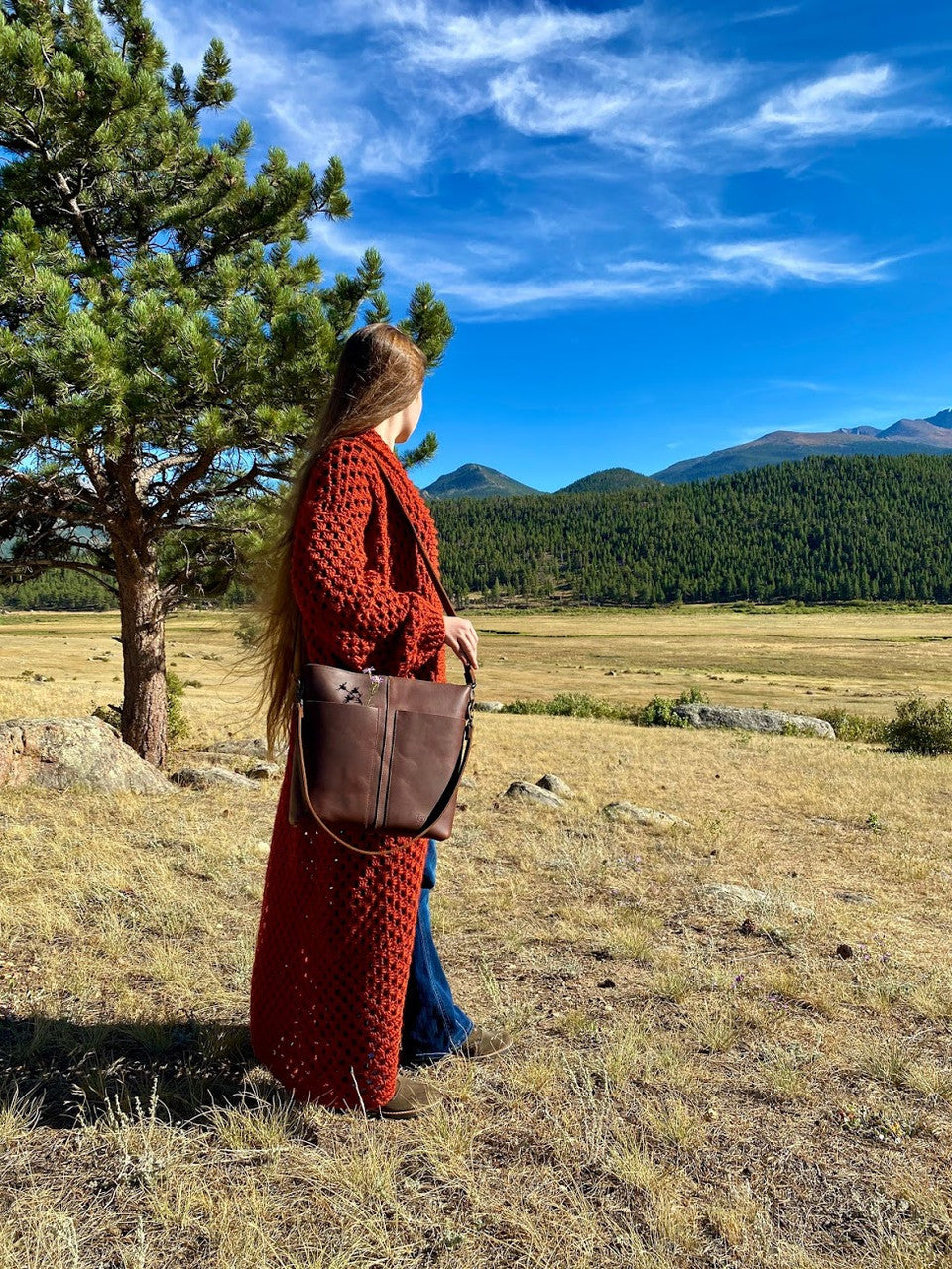 Minimal Leather Bag in dark brown, carried by a woman in a red knit coat in a mountain meadow