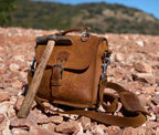 Indiana Leather Satchel in Tobacco on red rocky ground beside a wooden-handled rock hammer, hills behind
