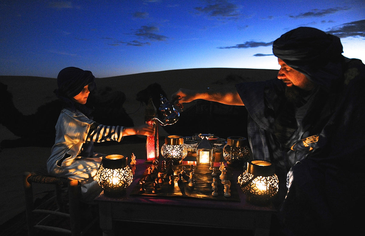Leather Travel Chess Set with pieces on a lantern-lit table in the desert at dusk as two people share tea