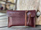 Cash & Receipt Bag in Chestnut, closed with leather tie, propped against a weathered tree stump outdoors