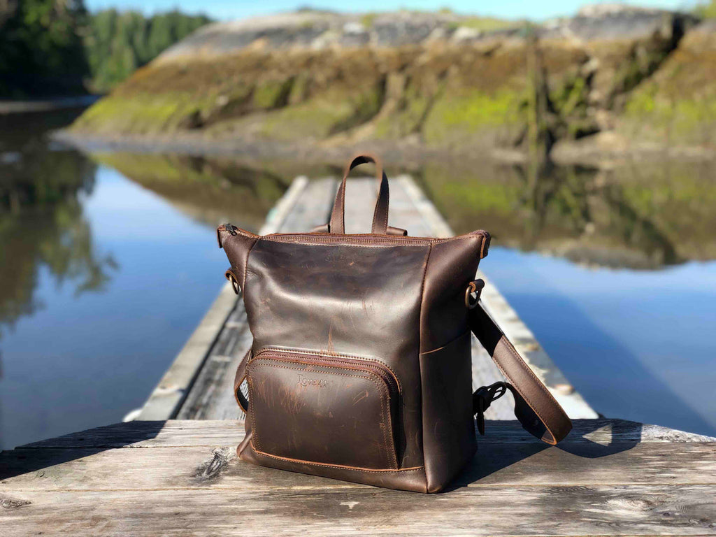 Universal Leather Backpack in Dark Coffee Brown, front view on a lakeside dock with hills reflected