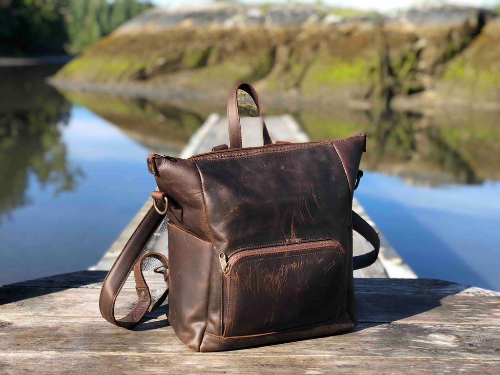 Universal Leather Backpack in Dark Coffee Brown, angled view on a wooden dock over a lake