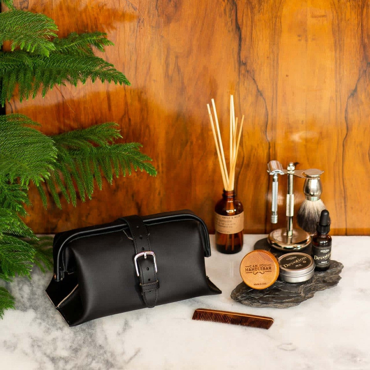 Big Mouth Leather Toiletry Bag in Black on a marble counter with a comb, shaving set and diffuser