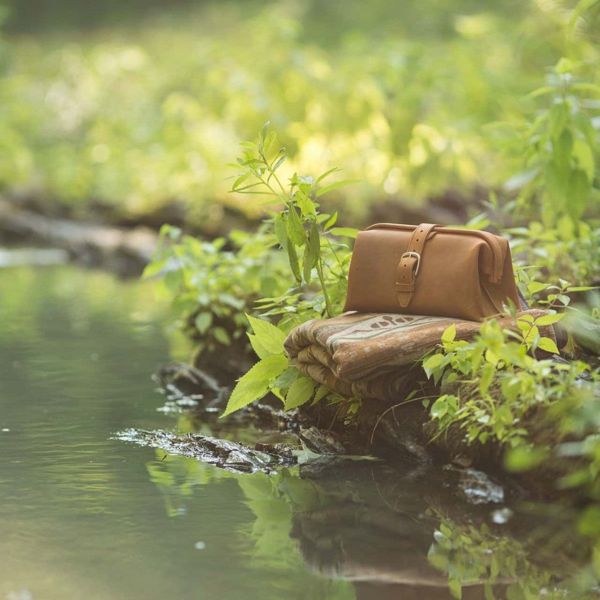 Big Mouth Leather Toiletry Bag in Tobacco on a folded blanket beside a creek surrounded by green plants