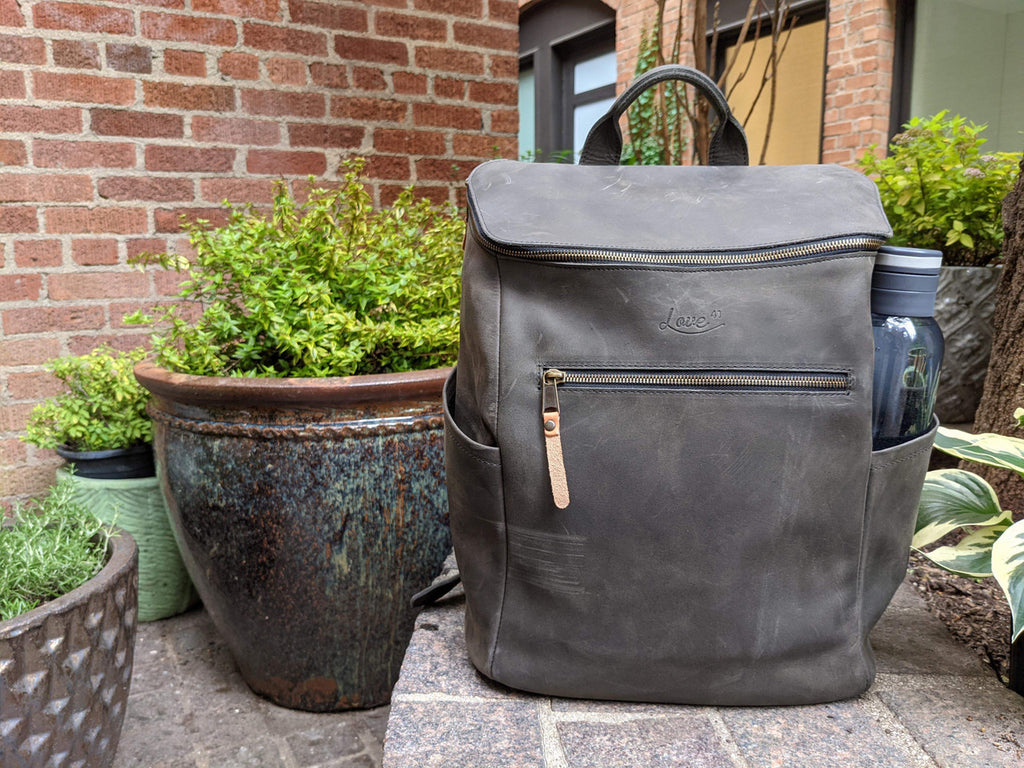 All in One Leather Backpack in Black on a brick patio beside potted plants with a bottle in the side pocket
