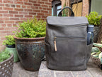 All in One Leather Backpack in Black on a brick patio beside potted plants with a bottle in the side pocket