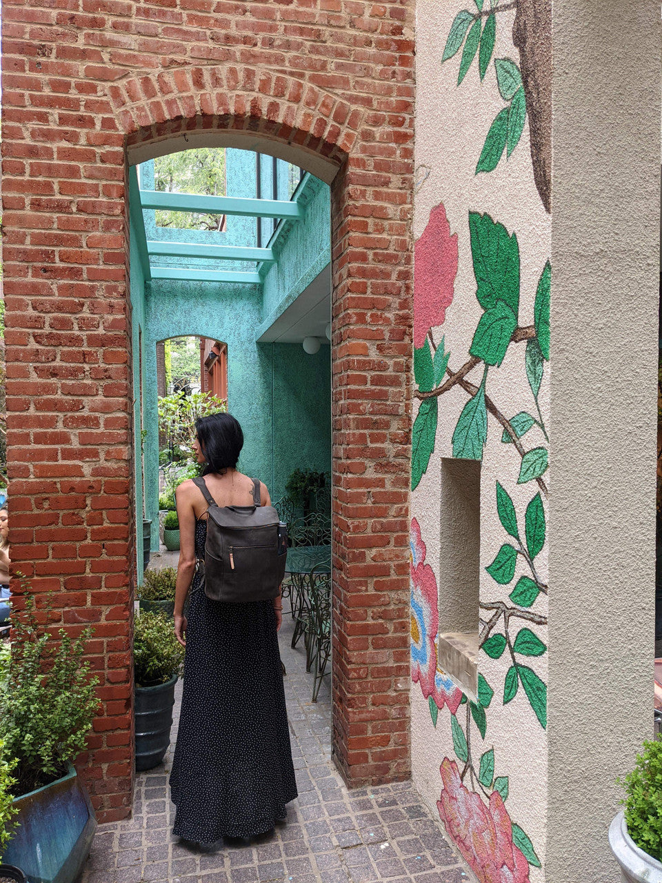 All in One Leather Backpack in Black worn by a woman walking through a brick archway beside a floral mural