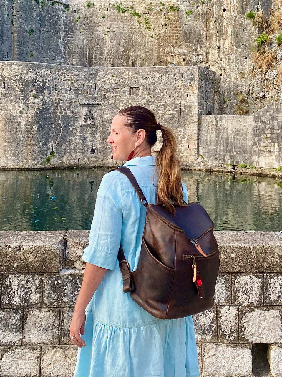 All in One Leather Backpack in Dark Coffee Brown worn by a woman in a blue dress beside old stone walls