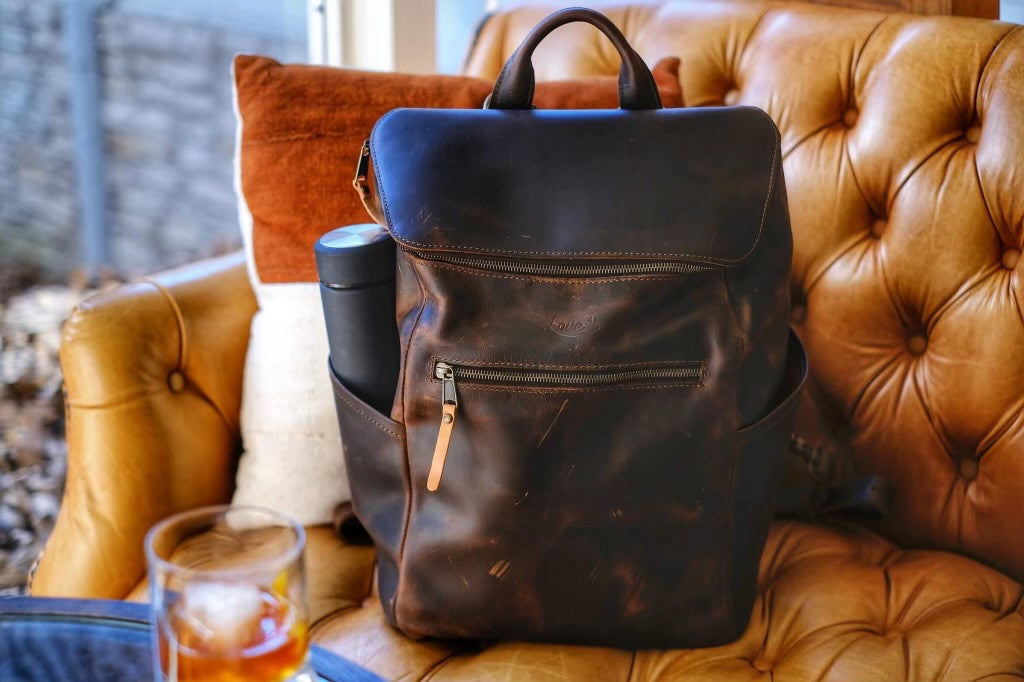All in One Leather Backpack in Dark Coffee Brown on a tufted leather armchair beside a glass on ice