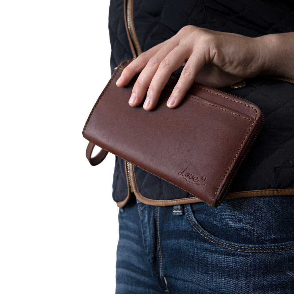 Medium Wristlet Leather Wallet in burgundy brown, held against a quilted vest and jeans