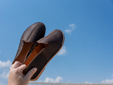Hand holding up a pair of Barefoot Slip-on Shoes in Dark Coffee Brown against a blue sky