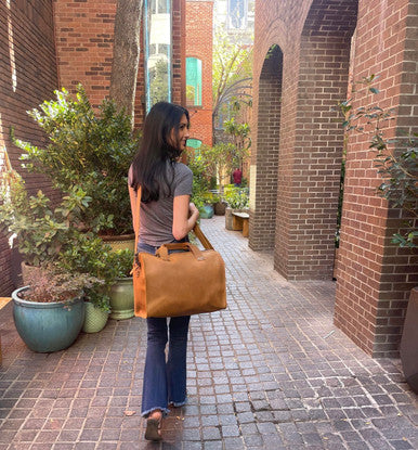 Simple Overnight Leather Duffle Bag carried by a woman walking down a brick alley lined with plants
