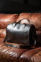 Simple Overnight Leather Duffle Bag in Dark Coffee Brown, resting on a worn brown leather sofa