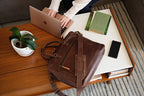 Women's Business Satchel in Chestnut, lying on a coffee table while a woman types on a laptop