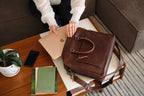 Women's Business Satchel in Chestnut, resting on a coffee table beside a laptop, books and phone