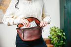 Leather Tumbled Fanny Bag in Chestnut, unzipped at the waist as a woman tucks cash inside