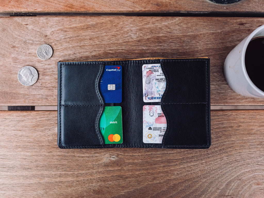 Large Bifold Wallet in Raven Black lying open with cards in the slots, beside coins and a coffee mug