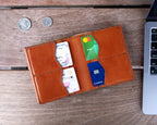 Large Bifold Wallet in English Tan lying open with cards in the slots, beside coins and a laptop