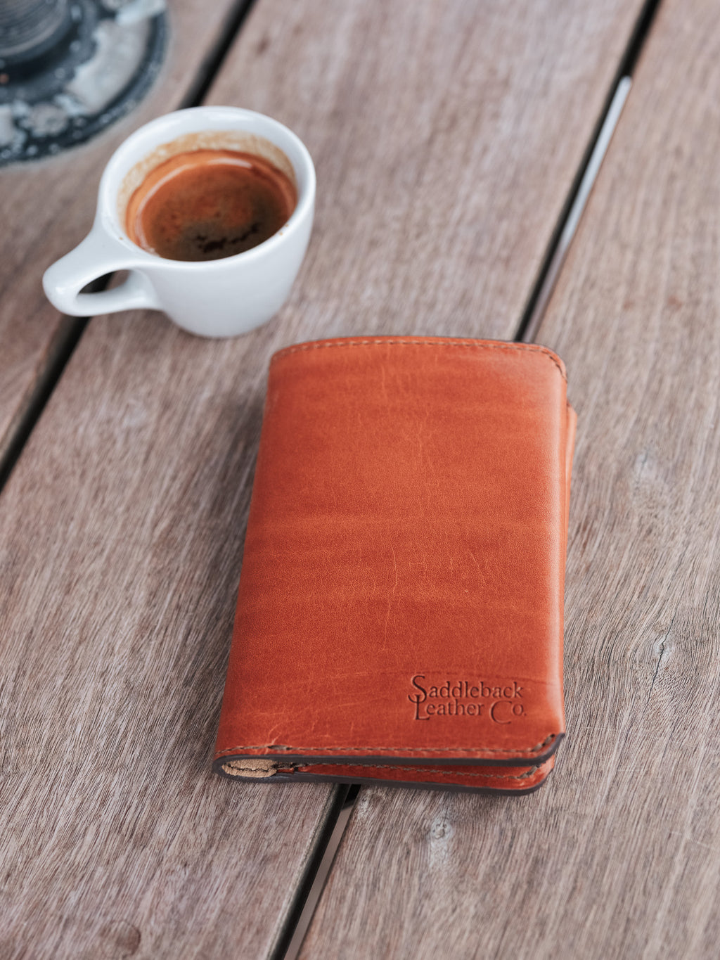 Wycliffe Passport Wallet in English Tan, closed with embossed logo, on a weathered table beside a coffee cup