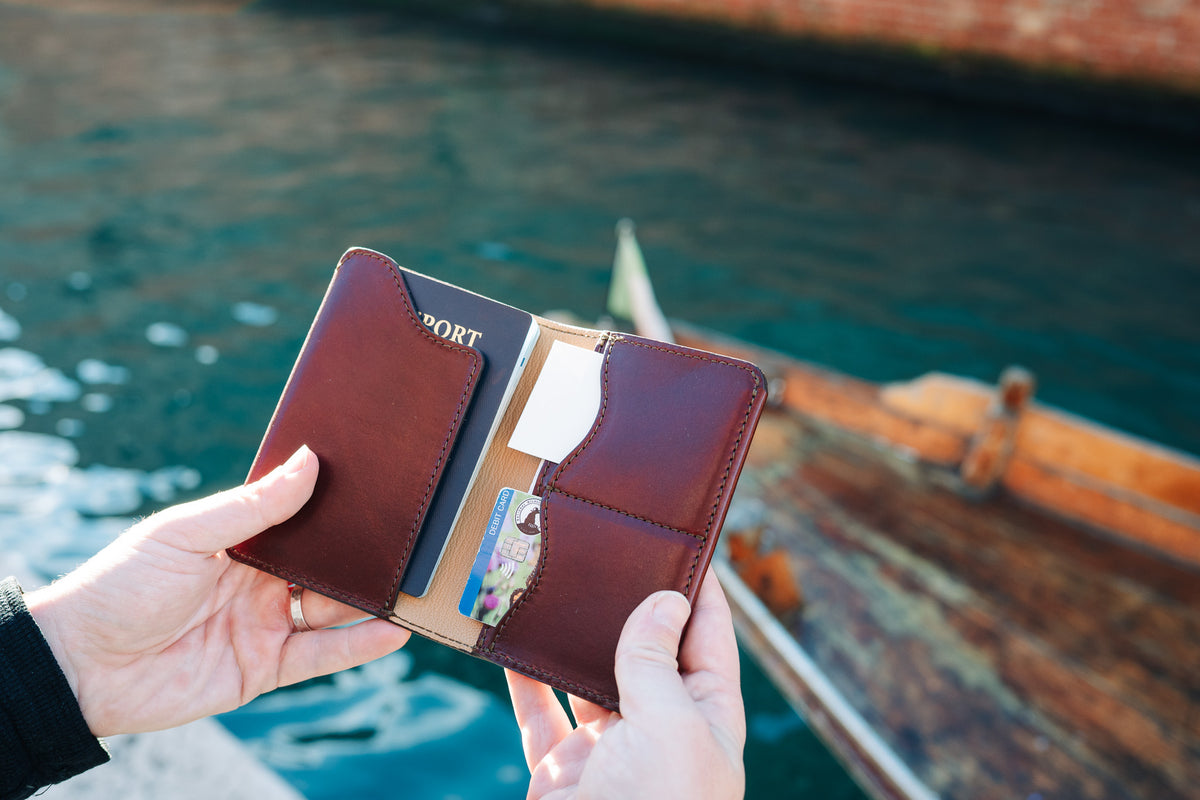 Wycliffe Passport Wallet in Oxblood held open in two hands with a passport and cards, by the water near a boat