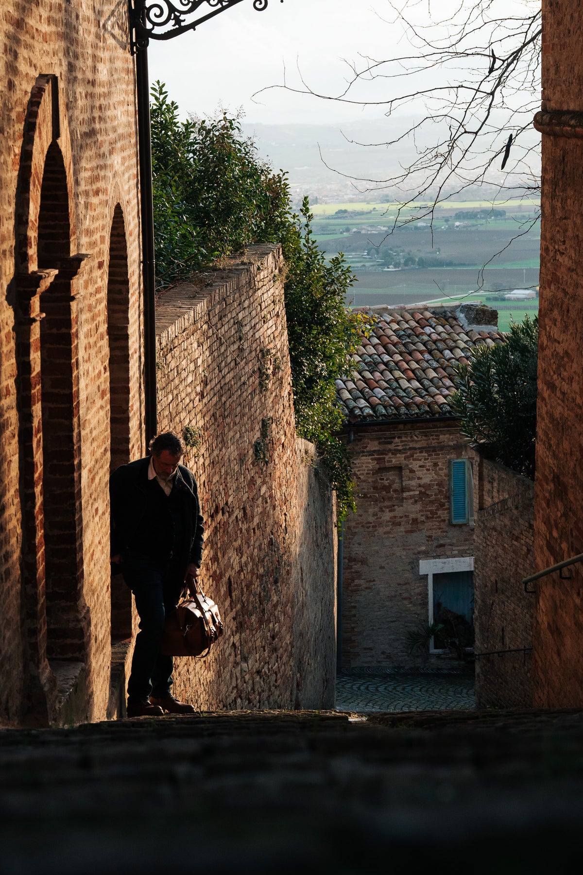 Wesley Deep Pocket Duffle in Raven Black, carried down a narrow brick alley in a hillside village