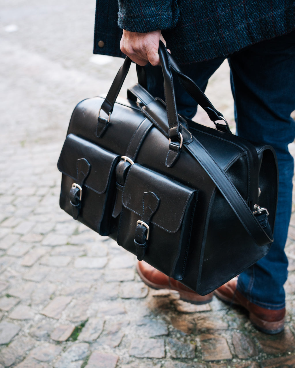 Wesley Deep Pocket Duffle in Raven Black, carried by the handles on a cobblestone street