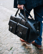 Wesley Deep Pocket Duffle in Raven Black, carried by the handles on a cobblestone street