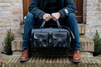 Wesley Deep Pocket Duffle in Raven Black, held by the handles while seated on brick steps