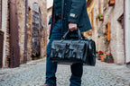 Wesley Deep Pocket Duffle in Raven Black, carried by the top handles in a village street