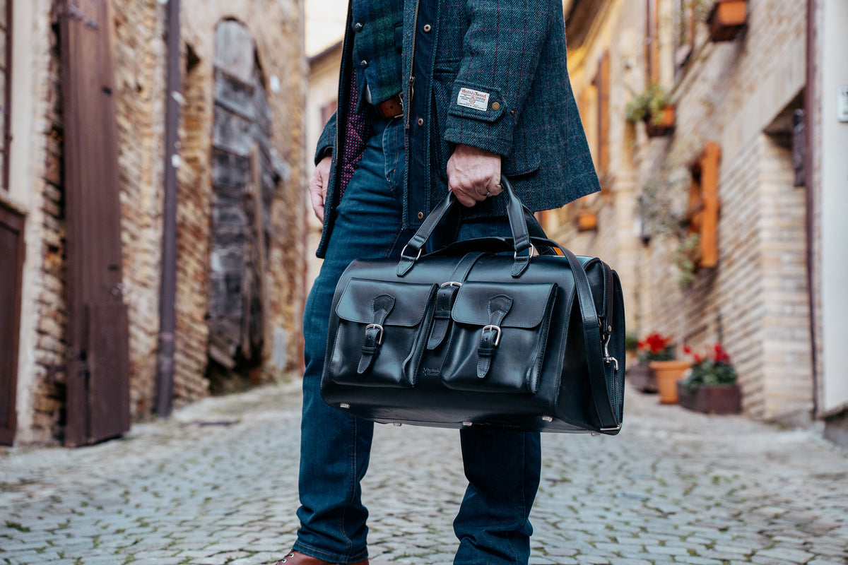 Wesley Deep Pocket Duffle in Raven Black, carried by the top handles in a village street