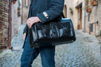 Wesley Deep Pocket Duffle in Raven Black, held at the side on a cobblestone street
