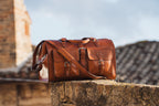 Wesley Deep Pocket Duffle in English Tan on a stone ledge with rooftops behind