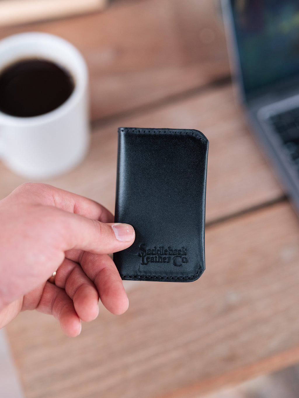 Slim Business Card Holder in Black, held in hand above a desk with coffee and a laptop