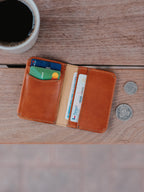 Slim Bifold Wallet in English Tan, open with cards on a wooden table beside coffee and coins