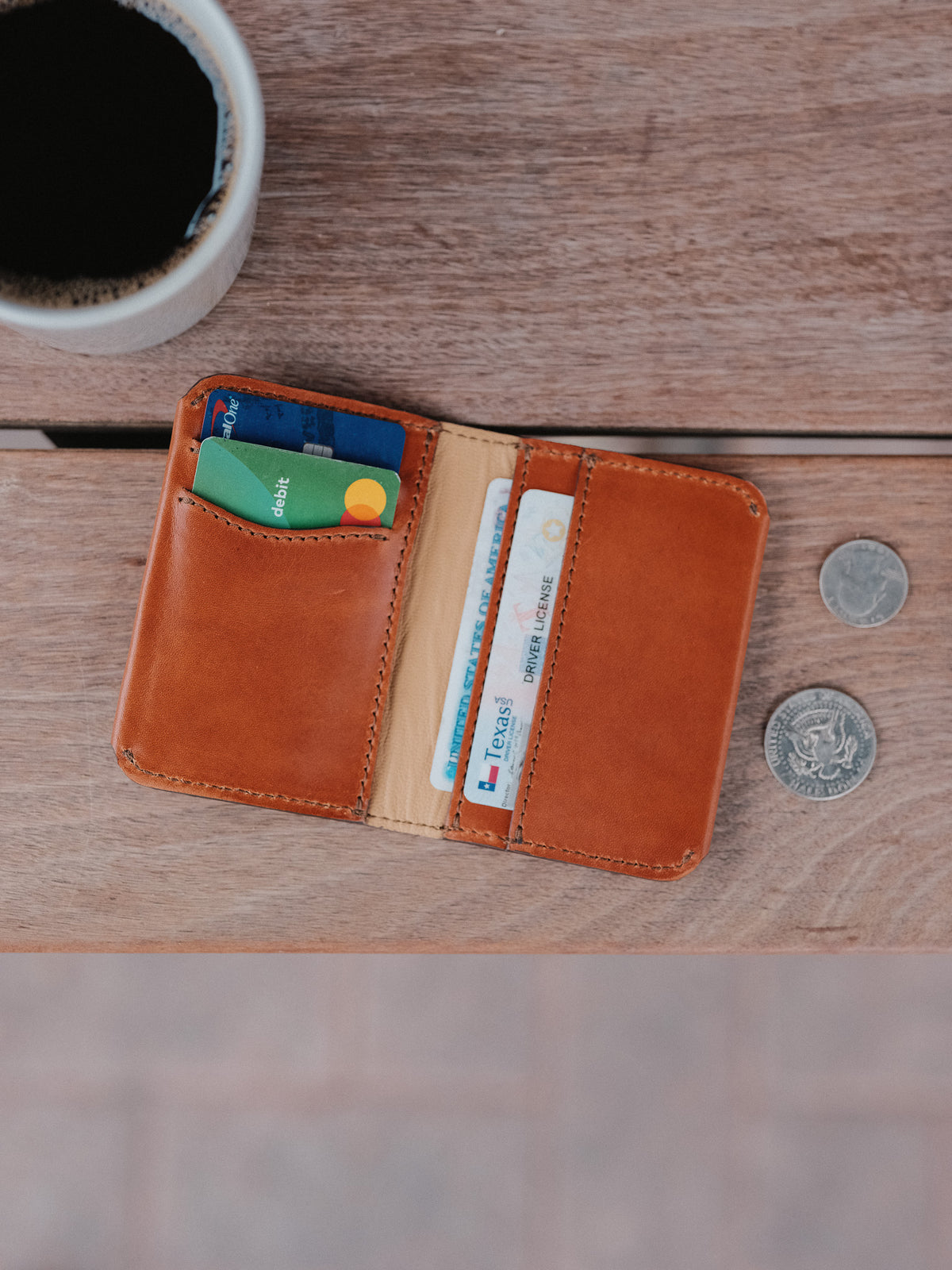 Slim Bifold Wallet in English Tan, open with cards on a wooden table beside coffee and coins