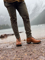 Huntsman Boots in Natural, worn with olive pants on a rocky lakeshore below misty mountains