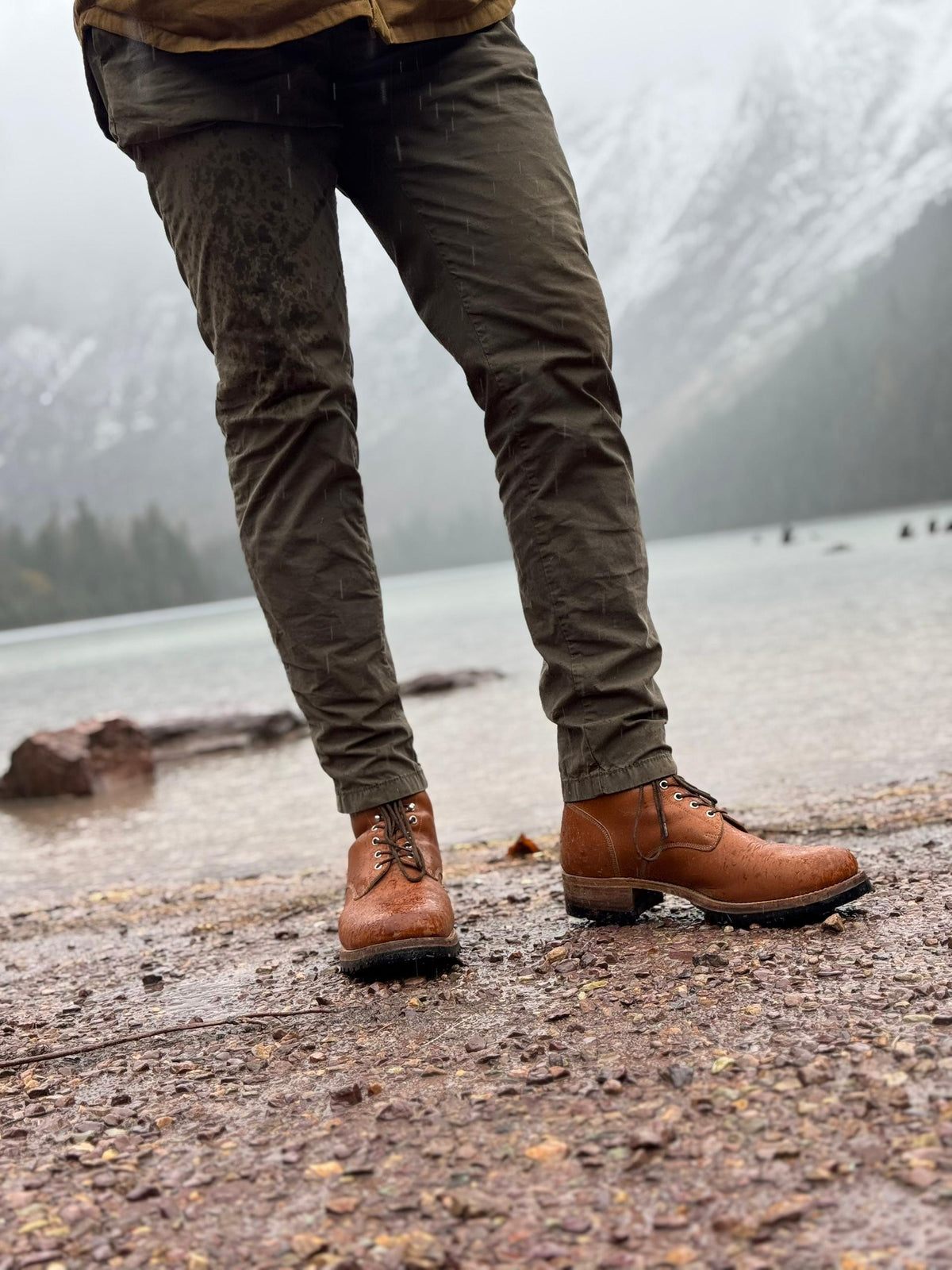 Huntsman Boots in Natural, worn with olive pants on a rocky lakeshore below misty mountains