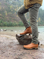 Huntsman Boots in Natural, one foot on a rock by a lake with forested hills behind