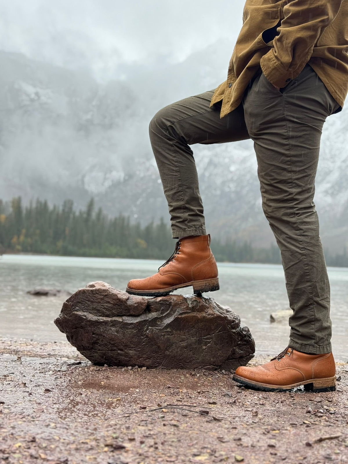 Huntsman Boots in Natural, one foot on a rock by a lake below misty mountains